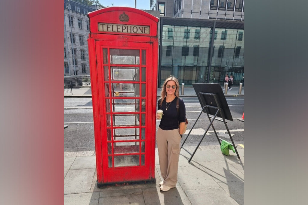 A woman in a black shirt stands next to red phone booth in London.