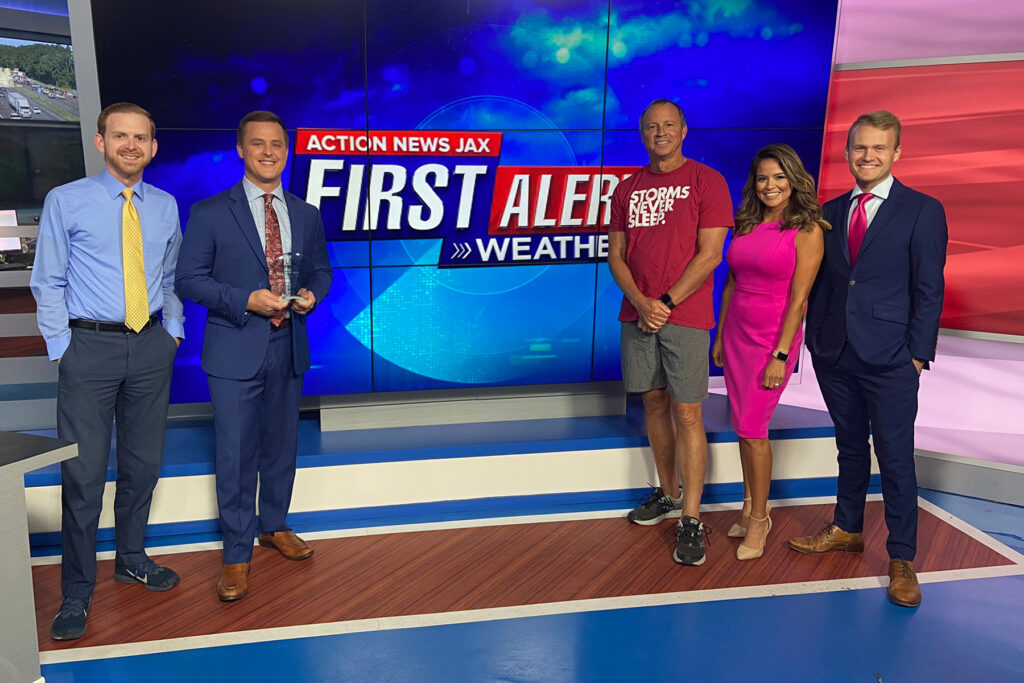 Woman in pink dress stands with other newscasters in front of a screen.