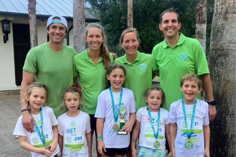 Four adult in green shirts stand behind four smiling children wearing medals.