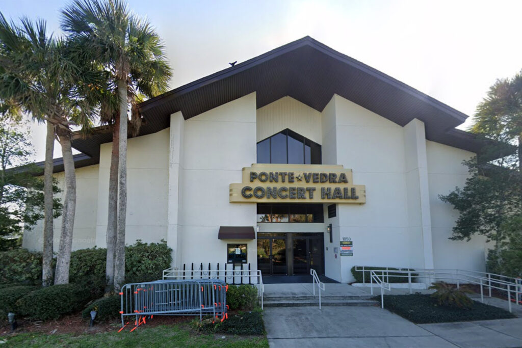 White building with a brown peaked roof flanked by palm trees.