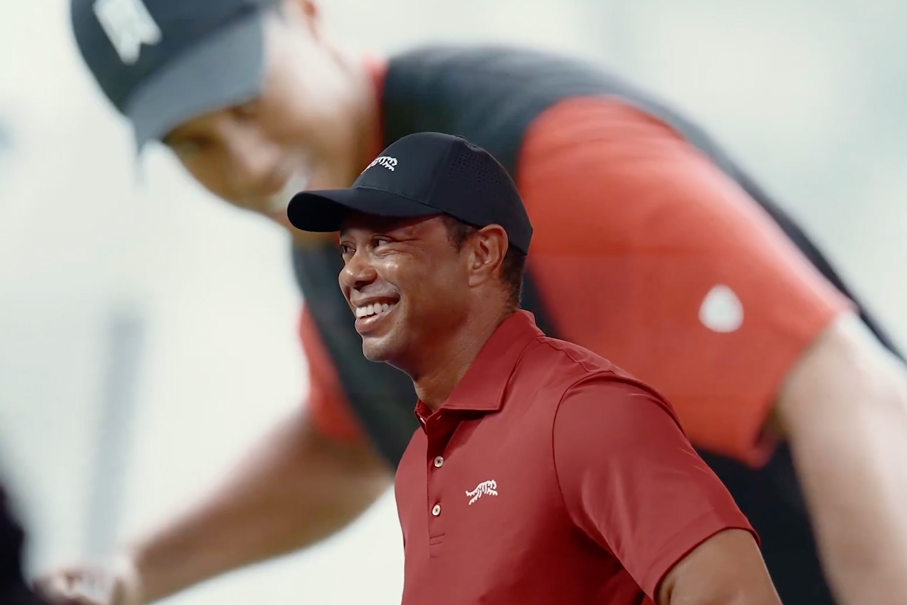 Tiger Woods wearing a red shirt stands in front of a large screen displaying an image of hi playing golf.