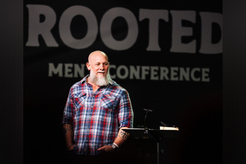 Pastor with white beard wearing a red-and-blue flannel shirt in front of a podium.