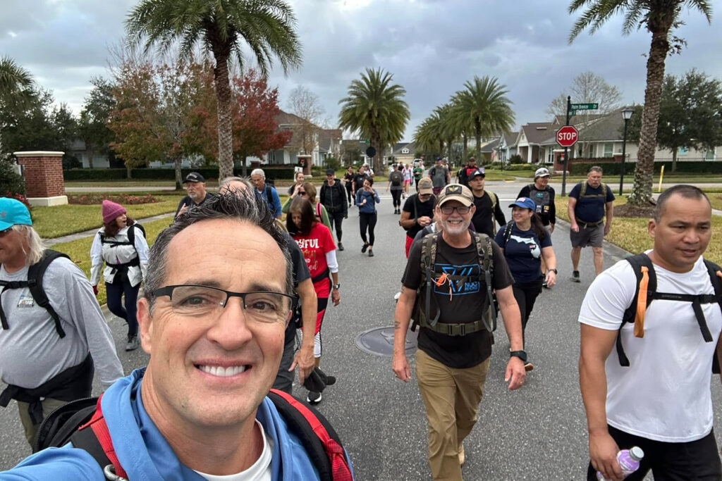 Man smiles outdoors with group of people wearing heavy backpacks. 