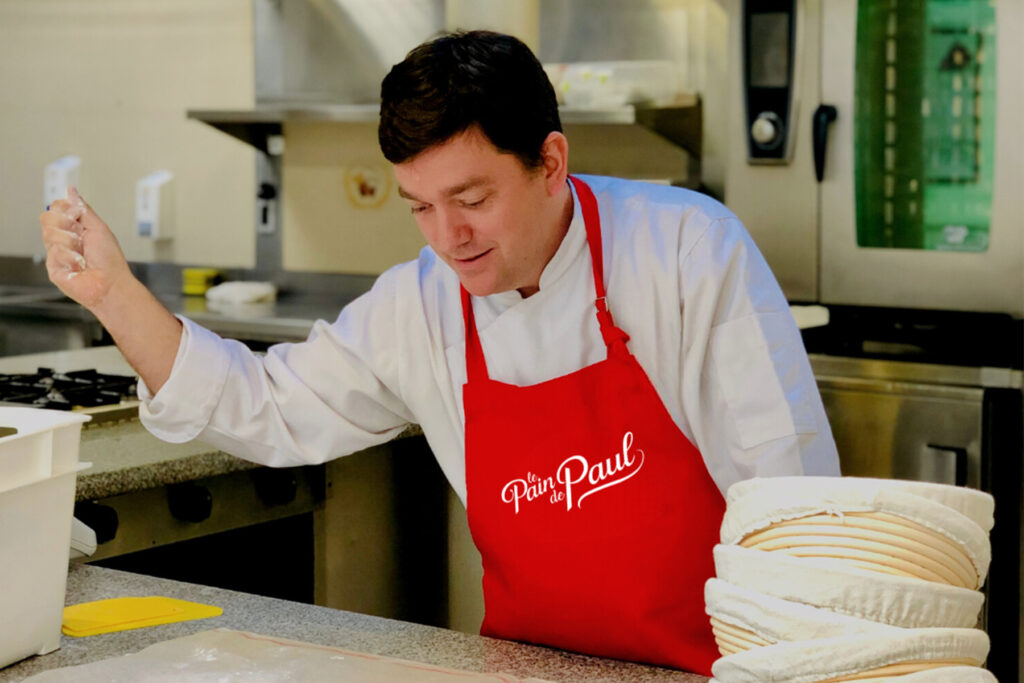 Man in red apron throws flour over dough.