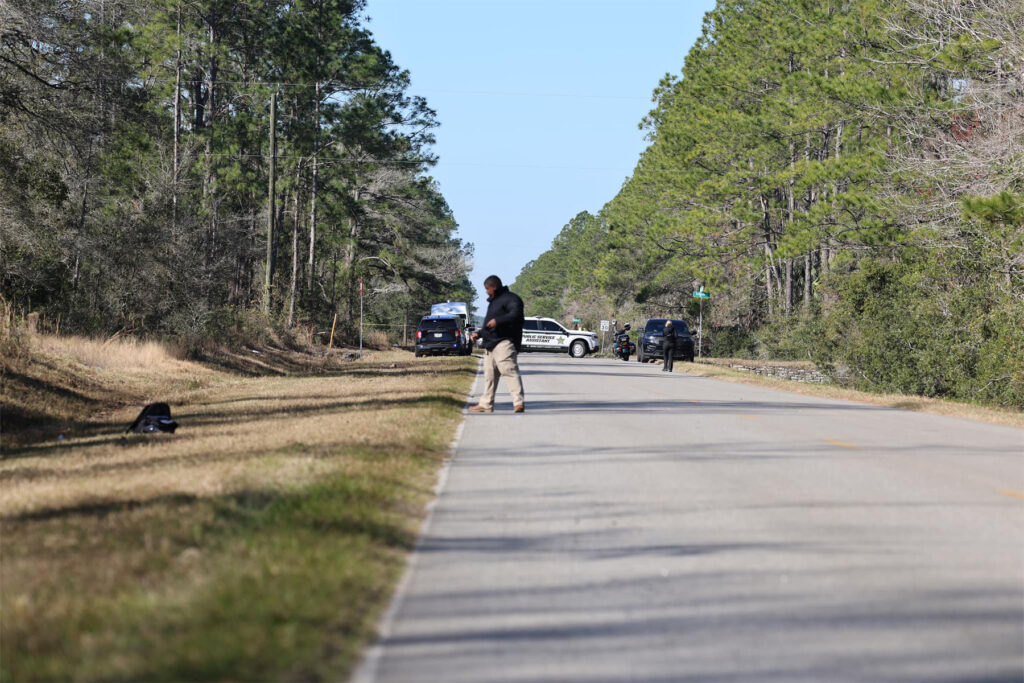 Investigator in middle of rural around way with police cars in the background.
